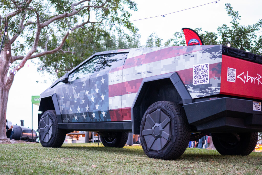 Rear-side angle of Cybertrucks with patriotic wrap and QR codes at a South Florida public event.