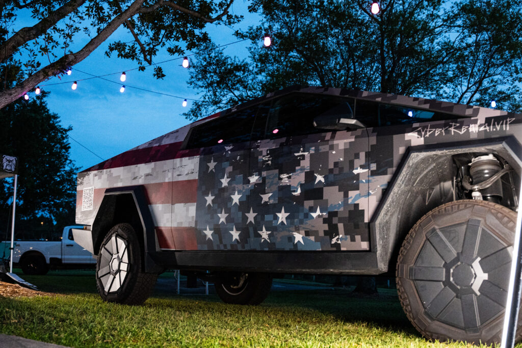 Cybertruck wrapped in American flag camo design displayed under string lights in South Florida evening event.